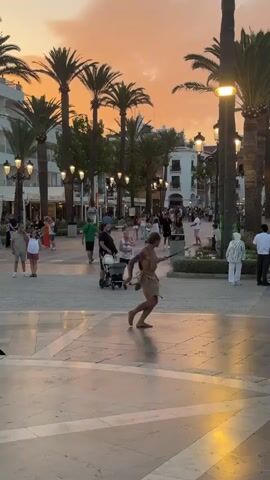 The video shows a public square during sunset. The sky is filled with warm, orange and pink hues, and palm trees line the sides of the square. Several people are walking around, some are sitting, and a few are standing in small groups. In the foreground, a man in a tan, loincloth-like garment is performing a dance or some kind of street performance with a stick. To his right, a young man in a blue and white athletic shirt and dark shorts is walking towards the camera, looking down at his phone. In the background, more people are visible strolling and congregating near buildings and shops. The ground is paved with light-colored tiles arranged in a geometric pattern. The overall atmosphere appears to be lively and casual, typical of an evening in a tourist area.