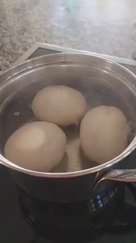 A top-down, close-up shot shows three light brown chicken eggs in a stainless steel pot of boiling water. The eggs are partially submerged and are being cooked on a black stovetop. One of the eggs has a visible crack on its surface. Steam rises from the boiling water.
