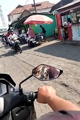 The video shows a street scene with several motorcycles and people. In the foreground, the perspective is from someone riding a motorcycle, with a hand and part of the handlebar visible in the bottom of the frame. In the middle ground, a dirt road is visible, with several motorcycles parked and moving. A person on a motorcycle wearing a helmet and a white shirt is on the left. Further back, a red food stall or shop is visible with people around it. On the right side of the stall, a person in a blue shirt and a light-colored head covering is seen. Other buildings with corrugated roofs are visible in the background. The lighting suggests it is daytime, possibly sunny. The overall impression is a busy street market or residential area with a lot of motorcycle activity.