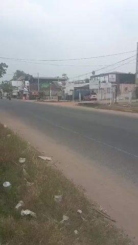 The video shows a moderately wide paved road with a dusty shoulder on the right side.  Grass and scattered debris are visible along the shoulder. Across the road, there are several buildings lining the street, some appearing to be shops or businesses, with awnings and signs.  There are a few vehicles present, including at least two motorcycles traveling on the road. The sky is overcast, giving the scene a muted and somewhat hazy appearance.  Power lines are visible overhead.