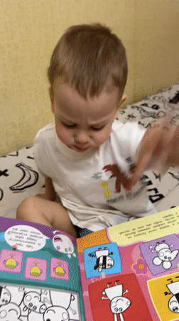 Toddler reads colorful book on bed in Rusanovo