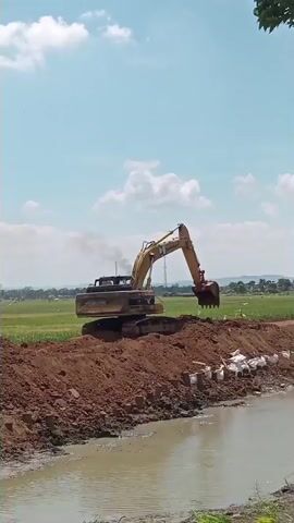 An excavator is digging dirt from a pile and moving it to a lower area next to a body of water. The excavator is yellow and has a long boom with a bucket. There is smoke coming from the exhaust of the excavator. The ground around the excavator is dirt and there are some white bags on the ground next to the dirt pile. The body of water is murky and appears to be a ditch or canal. In the background, there is a green field and a blue sky with white clouds. There are some trees visible in the upper right corner of the frame.