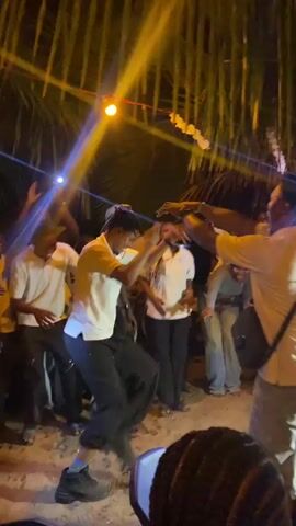 The video captures a joyous celebration with a group of people dancing together under string lights and what appears to be palm fronds. The two central figures are men dressed in light-colored shirts and darker pants, actively engaging in a lively dance. They are surrounded by a crowd of onlookers, many of whom are also dressed in light-colored clothing, cheering and recording the scene with their phone lights. The atmosphere is festive, filled with laughter and enthusiastic movement, with the background suggesting an outdoor evening event.