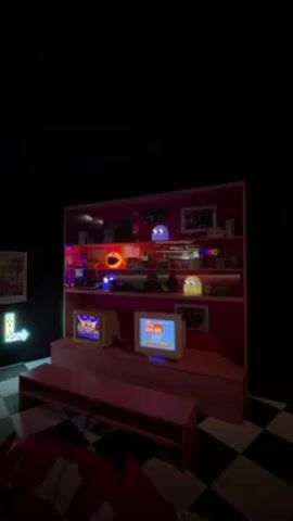 The video displays a retro-themed room with a prominent pink shelf filled with various items. Among them are glowing Pac-Man ghost figures and other nostalgic objects. Two vintage televisions sit on a pink platform below the shelf, both displaying animated graphics, including a possible Super Mario-style game. A checkered black and white floor adds to the retro aesthetic of the room. A red object, possibly a beanbag chair, is visible in the foreground. The environment is predominantly dark, highlighting the glowing elements of the display.