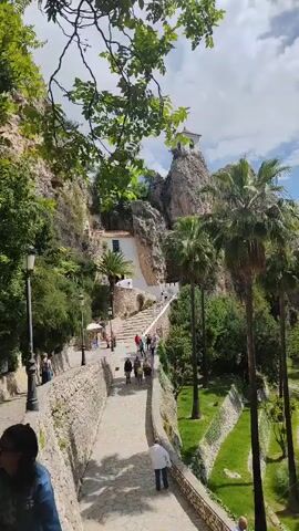 The video shows a view of a pathway leading to a building nestled within a rocky cliff. The path is lined with stone walls and leads uphill via a series of steps. Numerous people are walking along the path. Palm trees stand on the right side of the pathway, adding a tropical feel. A building with white walls and a small structure on top is perched atop the cliff, overlooking the scene.  The sky is partly cloudy, and tree branches frame the upper portion of the video. A woman in a blue denim jacket is partially visible on the left edge of the video. There are street lamps lining the path.