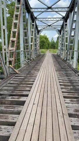 The video shows a view from an old, weathered wooden bridge. The bridge has a steel truss structure and the wooden planks of the walkway are aged and uneven. On the left side of the bridge, there is a section of railing with some blue paint chipped away. Beyond the bridge, a river flows through a lush green landscape with trees and undergrowth. The sky is partly cloudy, with white clouds against a blue sky. The camera pans slightly, providing a view of the river and the surrounding vegetation from the perspective of someone standing on the bridge.