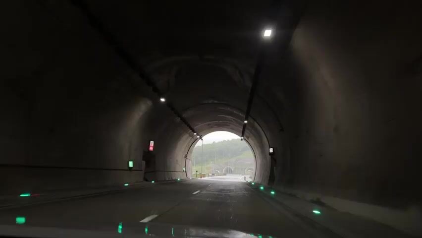 The video shows a car exiting a tunnel. The tunnel is lined with concrete and has overhead lights. The road is wet and there are green lights on either side. Outside the tunnel, the road stretches towards another tunnel in the distance. Street lights line the road. Green trees are visible on the hillsides. The sky is overcast. There is some rain visible on the windshield.