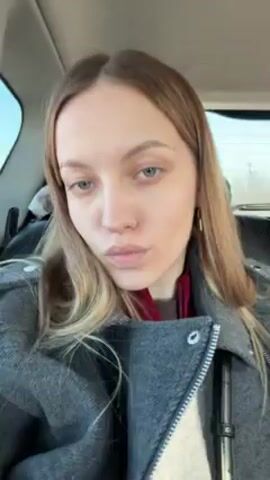 A woman with light brown hair is in the back seat of a car. She is wearing a gray coat and a red scarf. The car appears to be moving, and the sun is shining brightly. The woman looks directly at the camera with a neutral expression. There are rosary beads hanging from the car's ceiling.
