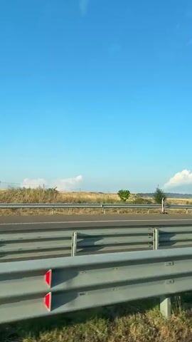 The video is a POV shot from a vehicle traveling on a highway. The highway has a metal guardrail in the foreground and appears to be a multi-lane road. On the right side of the highway, there is a field with dry grass and what appears to be a large solar panel farm. The sky is a clear blue with a few white clouds in the distance. There is a road sign with red and white markings indicating a curve.