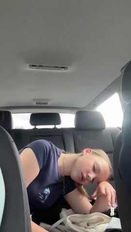 The video shows a young person sleeping in the back seat of a car. The person is leaning their head on their hand, with their eyes closed and mouth slightly open. They are wearing a dark blue t-shirt and a gold necklace. There are car seats visible in the foreground and background, and the car's interior lights are on. The lighting suggests it is daytime outside, but the view out the windows is not clear.