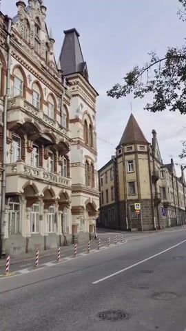 The video shows a street scene with several historic buildings on a cloudy day. The camera pans from right to left, focusing on the ornate architecture of the buildings. The building on the left is a large, multi-story structure with a light-colored facade, featuring brown decorative trim, arched windows, and balconies. The building on the right is also historic, with a brown and yellow facade, a conical tower, and arched windows. A road runs in front of the buildings, with white and red striped bollards lining the sidewalk. A pedestrian crossing sign is visible near the building on the right. The scene is quiet, with no people or vehicles visible.