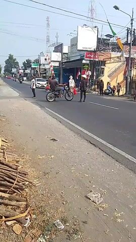 A street scene captures two men on motorcycles, facing each other and seemingly engaged in conversation or a transaction. The background reveals a row of shops, some adorned with Indonesian flags, indicating a local event or national celebration. Power lines crisscross overhead, and a few other vehicles and pedestrians are visible on the street. A pile of wood is stacked along the side of the road in the foreground, with some litter scattered nearby. The overall atmosphere appears to be that of a normal day with a touch of festive decoration.
