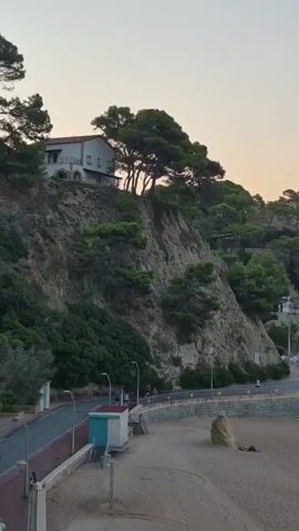 The video shows a cliffside with a white building on top and trees. Below the cliff is a beach with a road. There are a few people walking on the road. On the beach, there is a large rock and a person lying down. The sky is clear and light.
