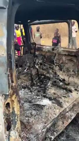 The video shows the inside of a completely burnt-out vehicle. The interior is charred and reduced to ash and twisted metal. Through the open frame of the vehicle, a group of people stand at a distance. The setting appears to be outdoors with some sparse vegetation in the background.