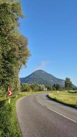 The video shows a scenic view of a winding road curving through a green landscape under a clear blue sky. On the left side of the road, there are tall trees with lush green foliage. Further down the road, a prominent mountain with a slightly rounded peak dominates the horizon. The mountain is covered in dark green vegetation, and a wisp of cloud or smoke can be seen near its summit. To the right of the road, a vast, sunlit field of green grass stretches out, dotted with a few scattered trees. A small building or barn with a red roof is visible in the distance within the field. The road itself is paved and has a white dashed line marking the center. Traffic signs are visible along the roadside, including a yellow directional sign with a curve and a red and white chevron sign warning of a bend. The overall impression is one of a peaceful, rural setting on a bright, sunny day.