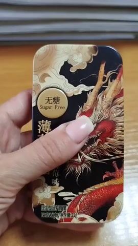 A person's hand is holding a small, rectangular tin with a black background and an intricate design of a red and gold dragon. The tin has a sliding plastic lid. The text "Sugar Free" is visible in a circular emblem on the front of the tin. The tin is being rotated slightly to show its different sides. The background appears to be a wooden surface.