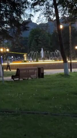 The video captures a park scene in Karachayevsk, Russia, likely during the evening or early morning. The lighting suggests it's dusk or dawn, with artificial lights illuminating the park. A prominent water fountain is the central focus, with multiple streams of water arching gracefully. Several people are visible walking around the fountain area and a path.

In the foreground, a wooden park bench sits on a grassy area. Adjacent to the bench is a trash receptacle. A lamppost with a glowing light stands to the right of the bench, with another lamppost visible further to the left. Trees with dark foliage frame the scene, and in the background, a mountain range with a distinctive rock formation can be seen, providing a strong geographical cue to the location. The overall mood of the video is tranquil and serene, emphasizing the beauty of the park with the interplay of water, light, and nature. No text is visible in the video.