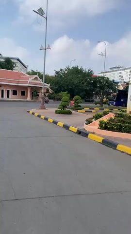 The video shows a street scene in Phnom Penh, Cambodia. The scene is framed by a road and sidewalk with a decorative roundabout containing a tall flagpole. Beyond this, a low building with a traditional Cambodian roof style can be seen, along with several parked cars and motorcycles. Three men are seated under umbrellas, appearing to be vendors or waiting for customers.  The sky is partly cloudy, and the overall atmosphere suggests a sunny day in an urban setting.