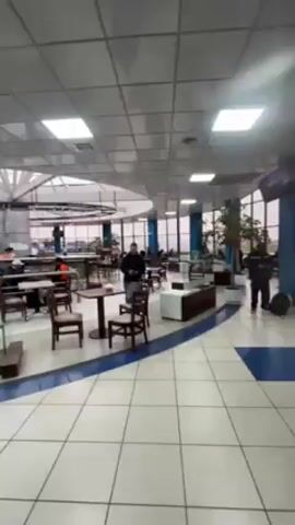 The video shows the interior of an airport. The first shot is an overview of a dining area with tables and chairs. An "Alexander coffee" sign is visible. The camera moves through a crowd of people, some with luggage. There are directional signs overhead. The environment appears to be an airport terminal. People are walking and talking, likely heading to their departure gates.