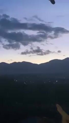 The video shows a landscape view of mountains and a city at dusk. The sky is a mix of light purple and gray, with clouds. A partially visible concrete pillar is on the right side of the screen. In the foreground, some vegetation is silhouetted. There are people sitting in the foreground, and only their feet and hands are partially visible. A water bottle is on the left edge of the screen.