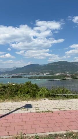 The video shows a scenic overlook with a paved walkway in the foreground. Beyond the walkway, there is lush greenery and then a body of water with mountains in the background. The sky is a clear blue with scattered white clouds. The video pans from left to right, capturing the expanse of the landscape. No people or animals are visible in the frame. The environment appears to be a coastal or lakeside area with a mountainous backdrop, likely on a sunny day.