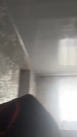 The video shows a room with a ceiling fixture, a window with curtains, and a wooden cabinet with glass doors. The camera is angled upwards, showing the ceiling and the top of the room. The lighting in the room appears to be dim. There are no people visible in the video. The scene is static and does not change throughout the brief clip.