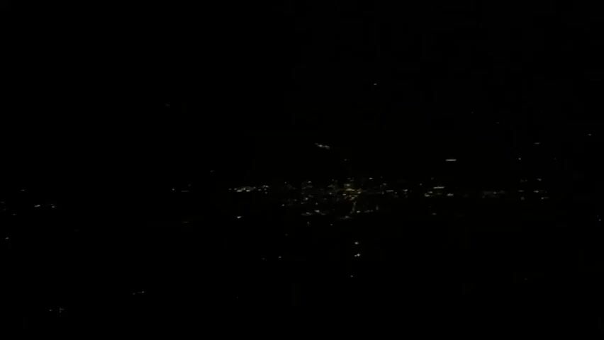 The video begins and ends with a black screen. In between, it shows a very dark, wide shot of a city skyline at night, viewed from a distance. Numerous small lights representing buildings and streetlights are visible, creating a faint glow across the horizon. The lights appear clustered in some areas and sparser in others, suggesting urban sprawl. There are no people, vehicles, or discernible objects in the frame, only the distant illuminated landscape against the darkness. The image quality is low, with grain and a lack of sharp detail, making specific identification of the location impossible.