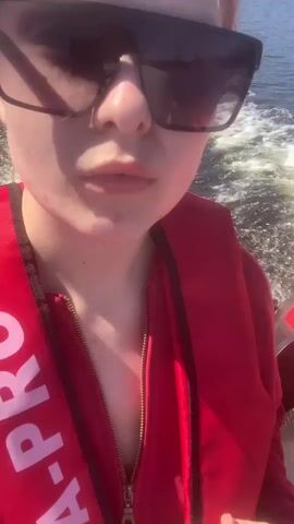 A person in a red life vest and sunglasses is seated on a boat, looking at the camera. The camera then pans to show the wake of the boat in the water, revealing a black outboard motor marked "9.9" at the stern. The boat is moving on a wide body of water, with a forested shoreline visible in the distance under a blue sky.
