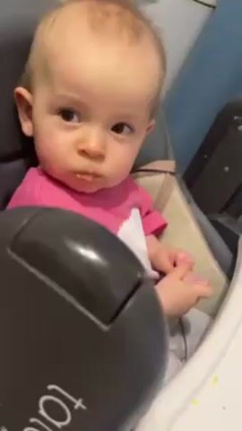 A baby sits in a highchair, looking around. The baby is fair-skinned with light brown hair and brown eyes, wearing a pink and white shirt. The highchair is gray and has the word "tatamia" on the front in white letters. The baby picks up a pink elephant toy and holds it in their right hand, looking at it briefly before looking away. The background shows a white cabinet, a light blue wall, and the edge of a dark gray appliance.