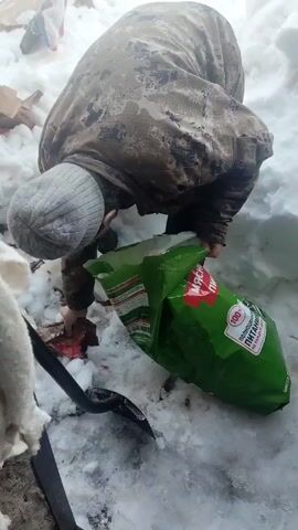 A man in a camouflage jacket and gray beanie is standing in the snow, emptying a green bag into a pile of trash. The bag is labeled with text in Russian. There is a black shovel and a pair of white gloves on the snow next to the bag. The man is bending over and appears to be struggling to empty the bag completely. The environment is snowy and appears to be an outdoor space with trash scattered around.