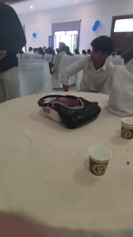 The video shows an indoor event, possibly a party or reception, with people seated at tables. A round table in the foreground is covered with a light-colored tablecloth. On the table, there is a black purse with red accents, and two paper cups with a gold design. A person in a black shirt and light-colored pants is standing near the table, holding a mobile phone. Other people are visible in the background, some seated at tables covered with white tablecloths and chairs draped in white fabric. Blue balloons are visible in the background. The lighting suggests an indoor setting, possibly a banquet hall.