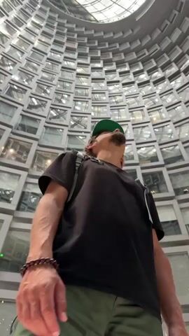 A man with a green baseball cap is standing in the center of a large interior space, gazing upwards. The man is wearing a black t-shirt and a backpack, and his head is tilted back to observe the architecture above. The space is a cylindrical atrium, lined with numerous rectangular windows arranged in a curved pattern, creating an eye-catching architectural design. A circular skylight is visible at the top of the structure, providing natural light to the area. The man is the central figure, his perspective emphasizing the height and complexity of the building's interior.