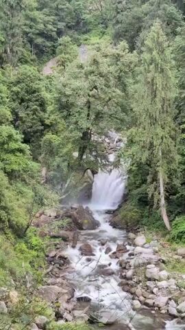 The video shows a waterfall cascading down a rocky mountain slope covered in lush green trees. The water flows into a rocky stream at the bottom. The sky above is partly cloudy.