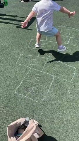 A young child wearing a pink hat, a striped t-shirt, and light blue shorts is playing hopscotch on a green, rubberized playground surface. The hopscotch grid is drawn with white chalk on the ground. The child is shown in motion, jumping between the squares of the hopscotch course. In the background, playground equipment is visible, including metal structures and a fence. A light-colored jacket or bag is lying on the ground in the foreground. The scene is lit by sunlight, casting shadows on the playground.