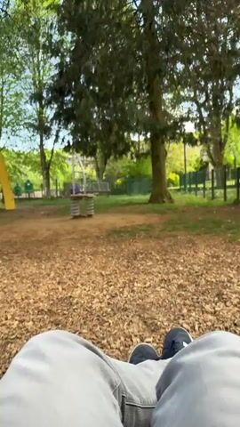 The video is filmed from a first-person perspective, showing a person's legs and feet as they swing. The person is wearing light grey pants and dark athletic shoes with white stripes. The view alternates between looking upwards at the sky and treetops and looking downwards at the ground in front of them.

When looking up, a clear blue sky with some clouds is visible, framed by the canopy of tall green trees. When looking down, the ground covered in wood chips is visible, typical of a playground surface. In the background, there are other trees, green grass, and playground equipment, including what appears to be a spring rider. The setting is an outdoor park or playground area in Pétange, Luxembourg, during daylight hours.
