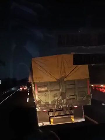 The video shows the rear of a truck at night, traveling on a highway. The truck is covered with a yellow tarp. In the distance, other vehicles can be seen with their headlights and taillights illuminated. The video appears to be filmed from inside another vehicle, with a person's silhouette visible in the foreground. The environment is dark, consistent with nighttime driving on a highway.