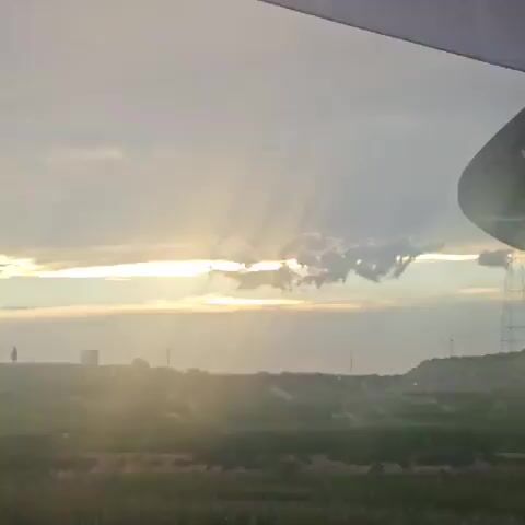 This video appears to be filmed from inside a vehicle, looking out through the windshield and side windows. The time of day is either sunrise or sunset, as evidenced by the warm glow of the sun breaking through the clouds and trees. The landscape outside the vehicle consists of green vegetation, possibly fields or hills, with some trees in the foreground and background. The sky is partly cloudy, with rays of sunlight visible. A rearview mirror is visible inside the vehicle, and a dashboard or part of the interior is also partially visible. The camera is in motion, giving a sense of travel, and the footage is somewhat blurred at times, possibly due to the vehicle's movement or camera focus. There are no people or distinct objects visible outside the vehicle other than the natural landscape. The overall atmosphere is serene and natural.