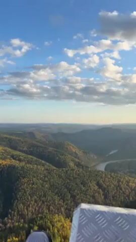 From a high vantage point, possibly a tower with a metal floor, the video captures a vast landscape of rolling, forested hills with a meandering river winding through the valley. The sky above is a bright blue, dotted with fluffy white clouds. There is also part of what seems to be the videographer’s foot in view.