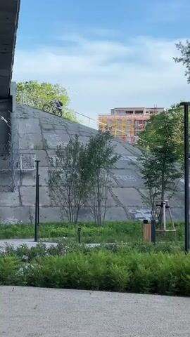 A cyclist rides a bicycle up a steep, concrete embankment. The embankment is covered in large, rectangular concrete slabs and has graffiti on its surface. A metal railing runs along the top of the embankment. In the foreground, a person walks their dog on a leash. Behind the embankment, a multi-story building with yellow and orange balconies is visible. The sky is blue with some clouds. The scene appears to be an urban park or recreational area.