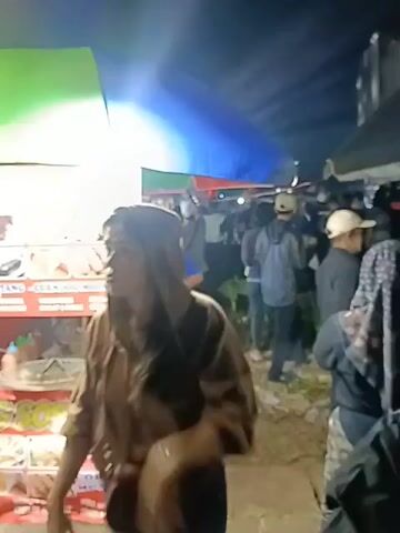 This video shows a street food stall selling corndogs and mozzarella hot dogs at night. The stall is decorated with colorful banners and has a menu board displaying various food items. Several people are gathered around the stall, some are customers, and others are vendors. The lighting suggests it's a lively night market or festival. The foreground shows a person wearing a head covering, partially obscuring their face, and the back of another person's head. The stall displays an assortment of fried food items, including corndogs on skewers. The overall atmosphere appears to be bustling and vibrant.
