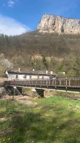 The video shows a serene view of a countryside landscape. A wooden bridge spans a shallow, rocky riverbed. The bridge is supported by concrete pillars and has a brown wooden railing. Several people are visible on the bridge, casually walking or standing near the railing. Adjacent to the bridge on one side is a white, single-story building with multiple windows, suggesting a residential or communal structure. The building has a gray roof. Behind the building and the bridge, a forested hill slopes upwards, culminating in a tall, rocky cliff formation. A cross can be seen at the very top of the cliff. The foreground features a grassy area with patches of purple wildflowers and occasional dandelions. The sky is blue, with scattered white clouds, indicating a bright, sunny day.