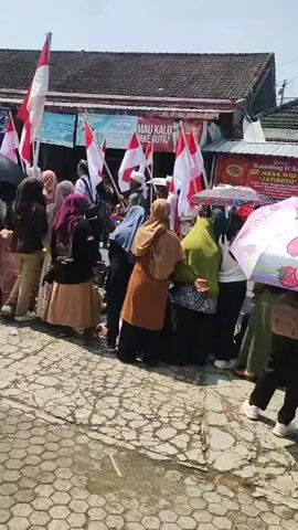 A group of people, predominantly women wearing hijabs, are gathered on a street. Many are holding Indonesian flags. The people are standing in a line, facing what appears to be a row of shops or stalls. One person is speaking into a microphone. The ground is paved with hexagonal stones. The day is bright and sunny.