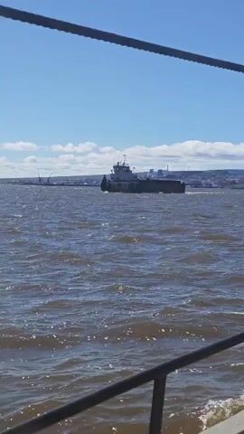 A barge is being pushed across a choppy body of water under a blue sky with scattered clouds. The barge is in the middle ground of the shot, and the foreground is occupied by the railing of a boat from which the video is being filmed. In the background, a city skyline is visible on the horizon. The video appears to have been filmed during the daytime, likely on a sunny day.