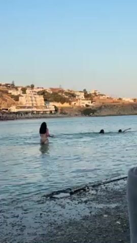The video shows a small bay at beach with three people in the water. Two are swimming further out, while the other, a woman with long dark hair wearing a bikini, is wading in closer to the shore. On the shore, there are some dark rocks and pebbles, and part of a gray sweatshirt sleeve is visible in the lower right corner of the frame, presumably of the person holding the camera. In the background, there are white buildings nestled into the hillside. The sky is a clear, light blue. It appears to be late afternoon or early evening, as the light is soft.