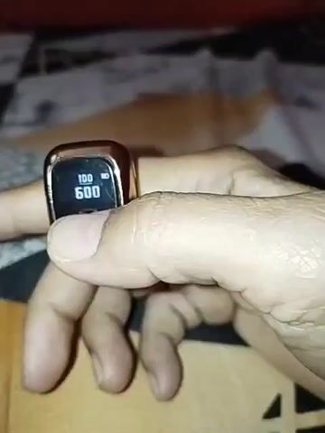 A close-up shot shows a person's finger wearing a rose gold colored smart ring. The ring has a small black digital screen displaying a battery icon and the number 601. The numbers on the screen change throughout the video, counting up from 601 to 611. The background is blurred but appears to be a patterned fabric.