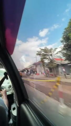 The video is filmed from inside a vehicle, looking out the passenger side window. The perspective suggests the vehicle is moving. Outside the window, there is a street with construction activity. A yellow caution tape runs along the side of the road, separating a construction area from the traffic lane. Several construction vehicles, including what appears to be a steamroller and an excavator, are visible. Workers in casual clothing are present, some engaged in tasks related to the construction. There are also houses and buildings visible in the background, suggesting a residential or mixed-use area. The sky is blue with scattered clouds, indicating daytime. The road appears to be under repair or construction, with loose soil and equipment present. Several motorcycles are also seen on the road, indicating ongoing traffic.