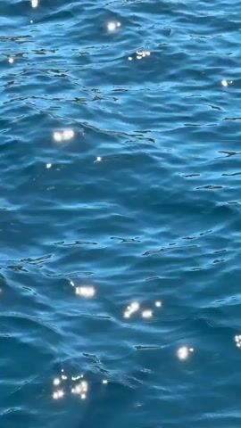 The video shows a close-up view of the surface of clear, turquoise-blue water with slight ripples. Sunlight reflects off the water, creating small, bright, flickering spots. The scene is static, and there are no visible objects or people other than the water itself and its reflections. Sounds of foreign speech can be heard.