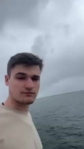 A young man stands on the deck of a ferry or similar boat, facing the camera. The background shows a body of water and a cloudy sky. As the camera moves, the architecture of the boat becomes more visible, including a white railing, windows, and a red-painted roof section. The light is flat and diffused, suggesting an overcast day.  The man's expression is neutral as he looks into the camera.