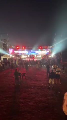 The video captures an outdoor nighttime event with a stage set up in the background. The stage features a large LED screen displaying the name "VAN NOSIKOV" in prominent white lettering, flanked by colorful, abstract graphics. Several bright spotlights are mounted above the screen, and red lights are positioned on either side of the stage. Two figures, presumably performers or DJs, are visible on the stage. A crowd of people is gathered in front of the stage, with some individuals dancing and others standing or sitting. Red lighting illuminates the ground in the foreground, creating a dramatic effect. The environment appears to be an open, unpaved area, possibly a field or a construction site, with some debris scattered on the ground. The overall atmosphere suggests a music or entertainment event.