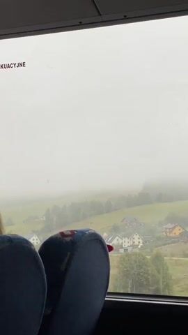 The video shows a view from inside a vehicle, looking out a window. The window has some text printed on it, which appears to be in a foreign language. In the foreground, two blue seats with some wear and tear are visible. Outside the window, a foggy, mountainous landscape is depicted. There are trees and houses scattered across the hills, suggesting a rural or small-town setting. The fog obscures the details of the background, creating a somewhat muted and hazy atmosphere.